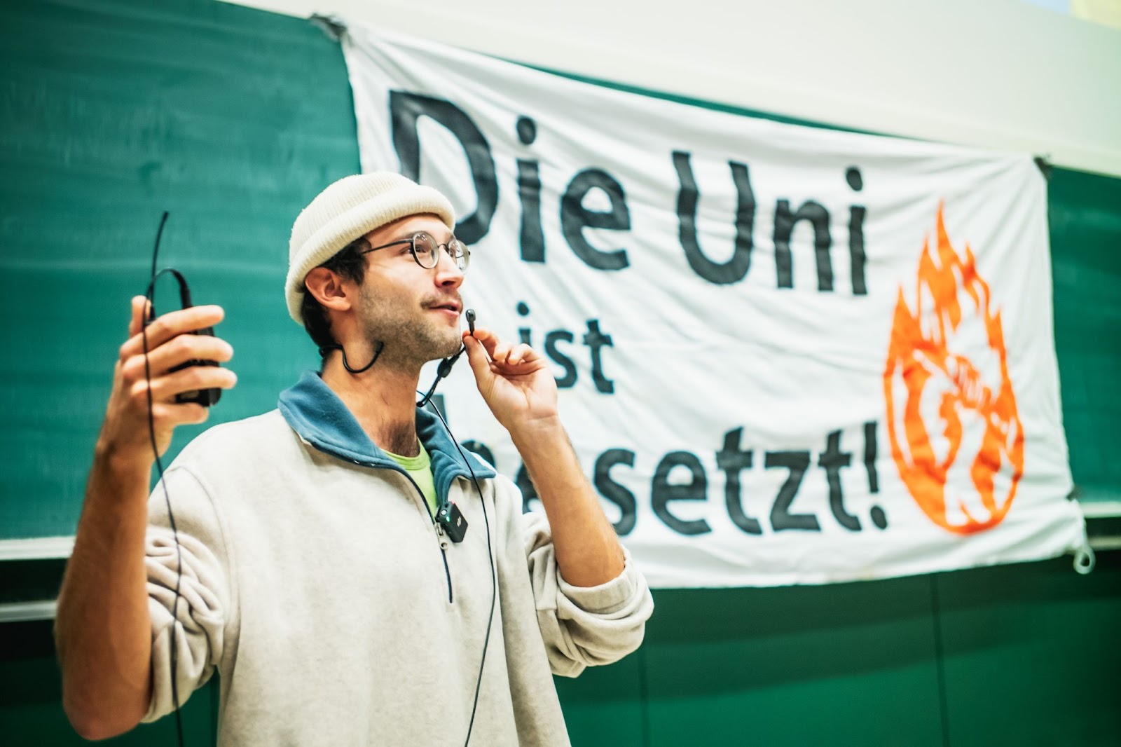 Ein Aktivist der Bewegung "Erde Brennt" in Wien spricht in ein Mikrofon. Im Hintergrund sieht man ein Plakat. In schwarzer Schrift steht auf weißem Hintergrund: "Die Uni ist besetzt!" Daneben das Logo von "Erde brennt": eine Flamme und darin eine zur Faust geballte Hand. Das Plakat hängt vor einer grünen Schultafel.