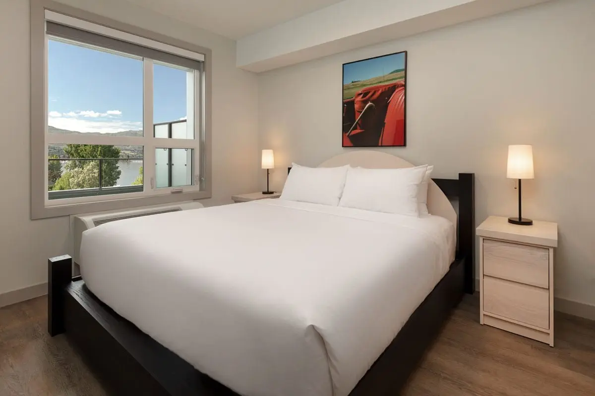 a one bedroom suite at The Shore Kelowna, a great option when booking kelowna hotels; room features a bed with white linen, two nightstands on either side with two lights turned on.