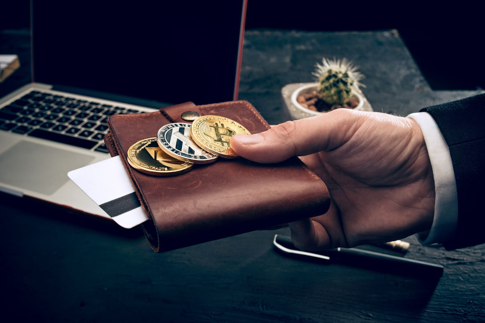 A men's hand is holding a wallet with crypto coins and payment card. Behind there is a laptop.
