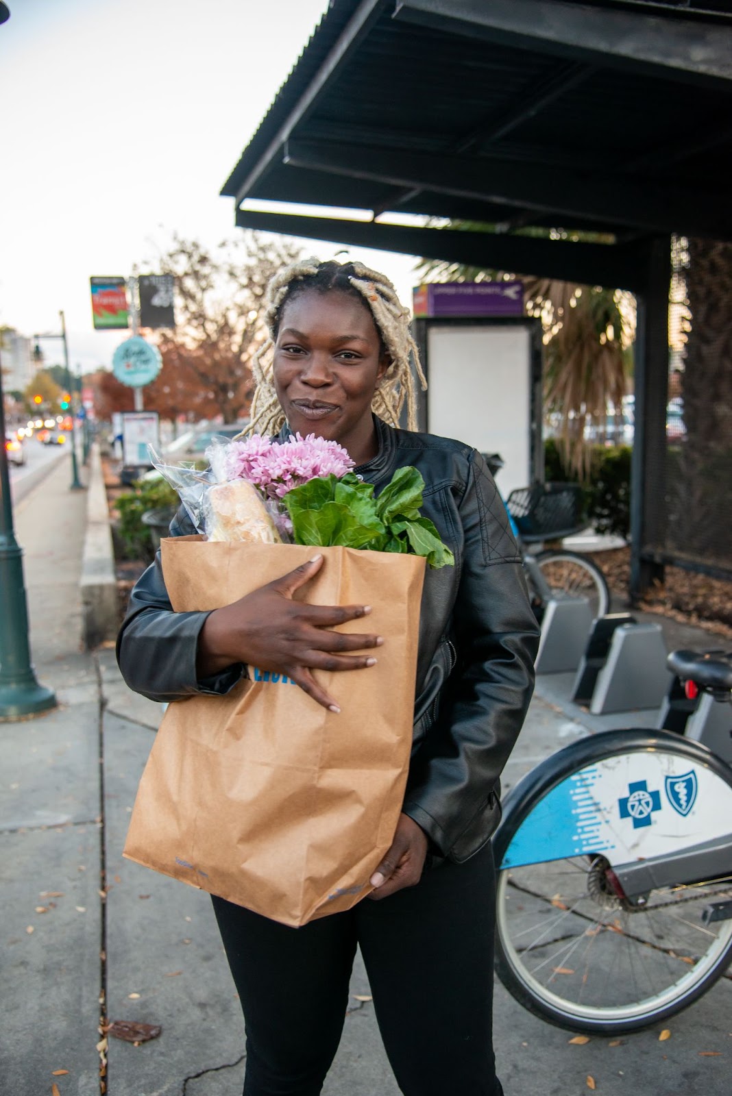 [A woman holding a bag of groceries that has bread and fresh flowers inside. The woman is standing near a COMET bus stop and Blue Bike SC location. The woman is black with blond braided hair and a leather jacket. She is smiling gently at the camera. ]