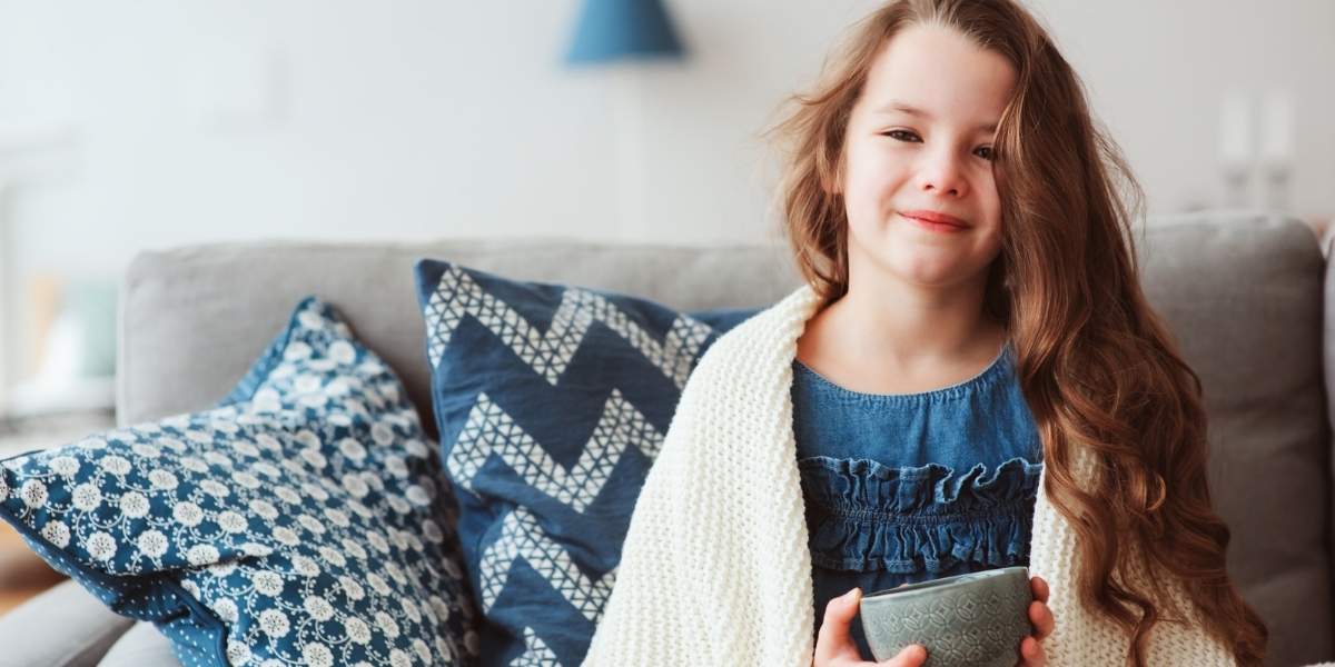 young girl wrapped in blanket on couch with tea smiling