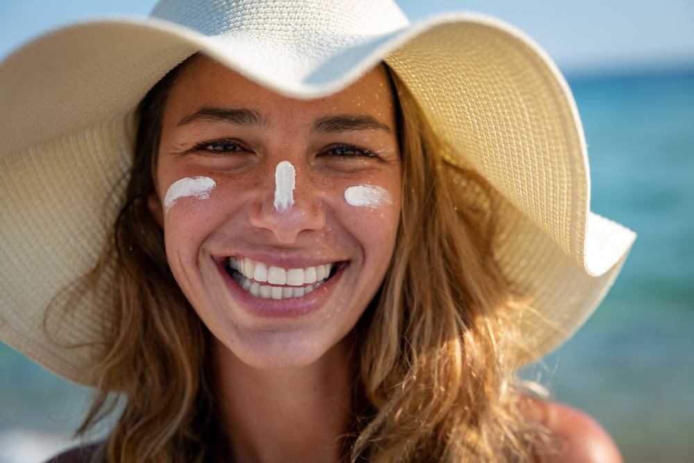 Close-up de uma mulher loira de cabelos compridos e usando um chapéu de palha sorrindo para a câmera com desenhos de protetor solar em seu rosto.