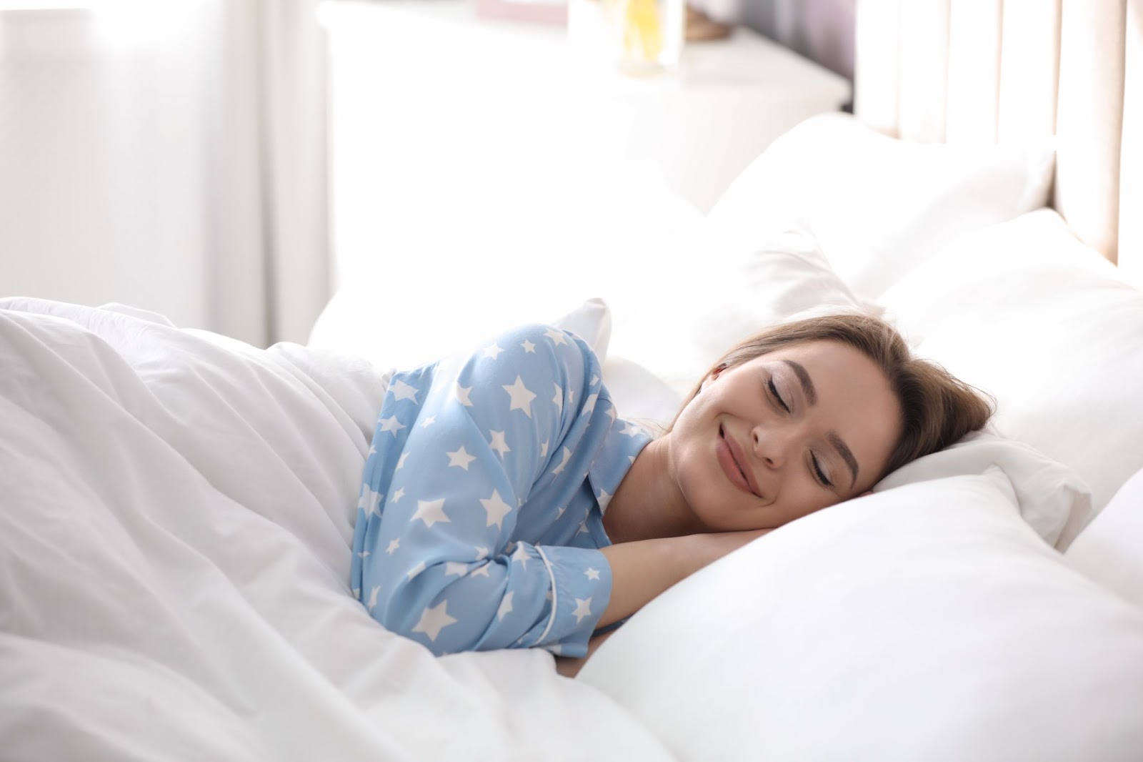 A woman lying in bed in blue pyjamas smiling with her eyes closed. 