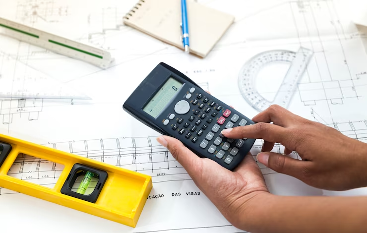 A woman using a calculator near a plan and engineering equipment.