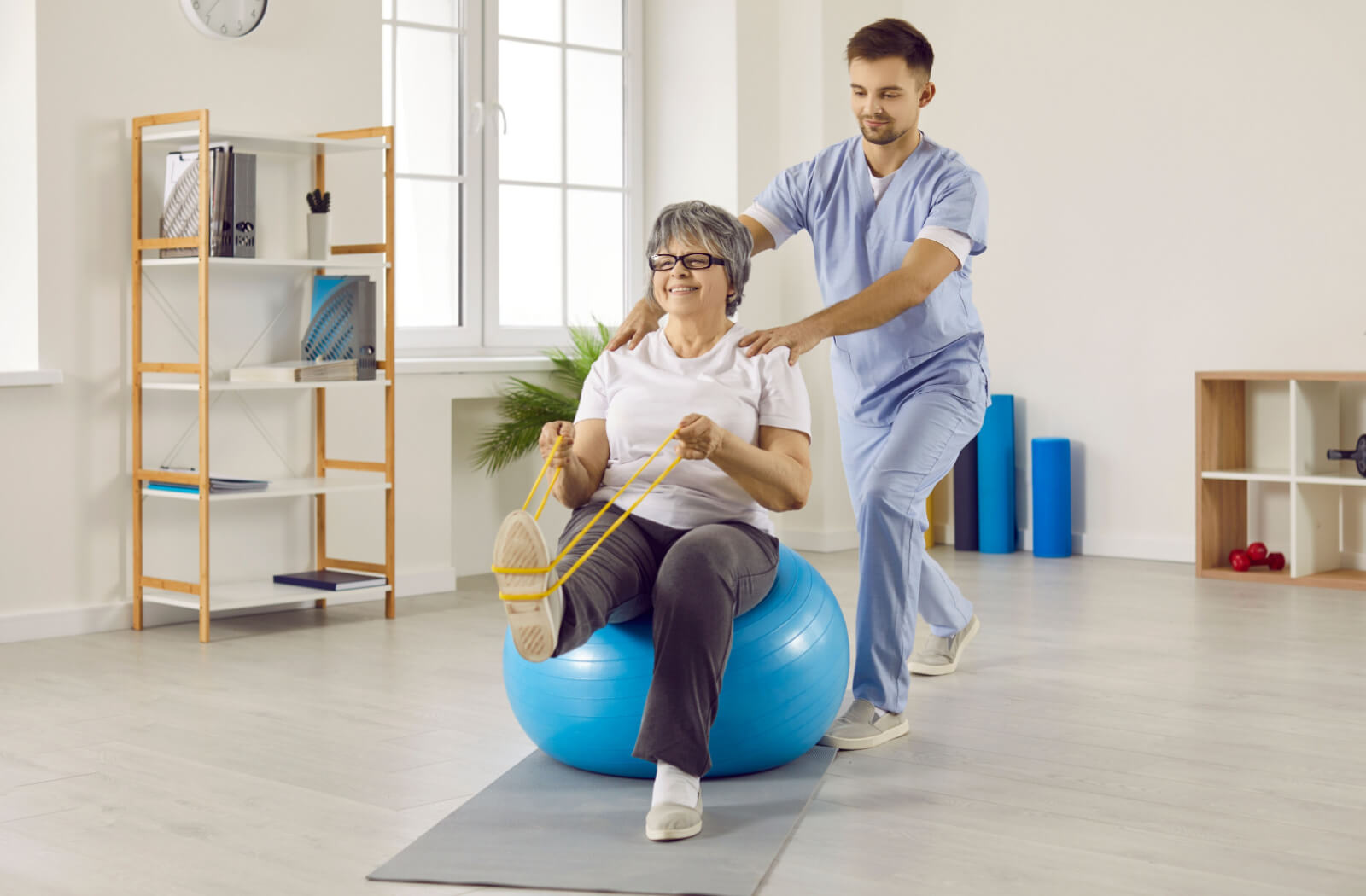 A smiling senior woman sitting on a fitness ball and doing exercise with resistance band, while being assisted by a nurse.