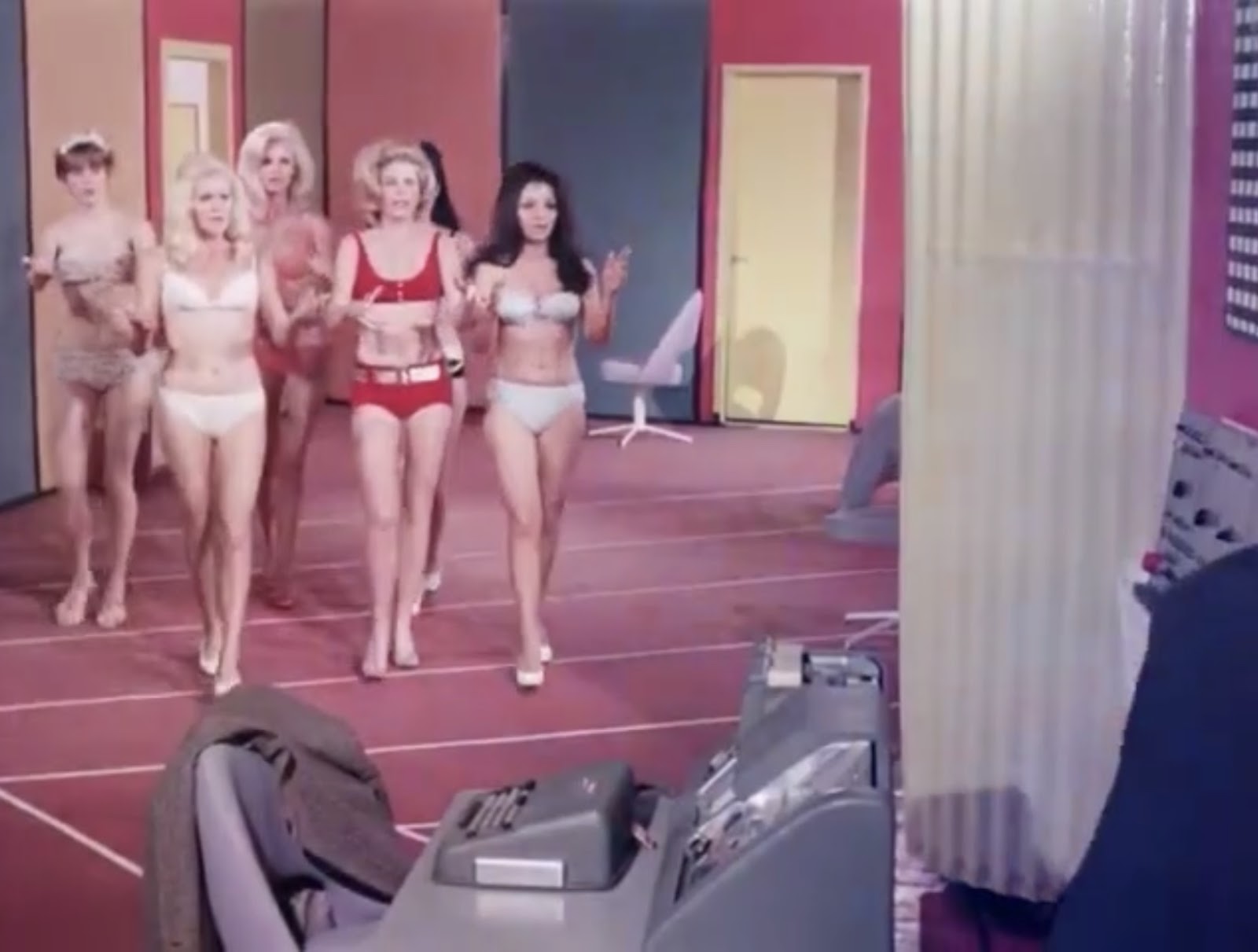 A still from How to Make a Doll. Six fembots wearing underwear walk in a brightly colored room.