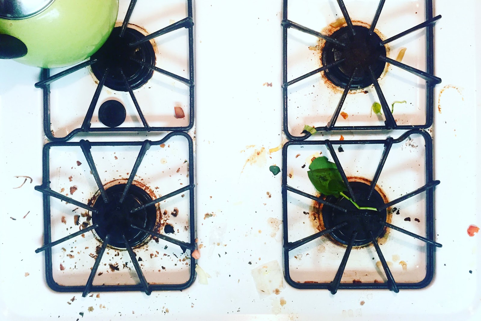 A gas stovetop with food crumbs, green teapot