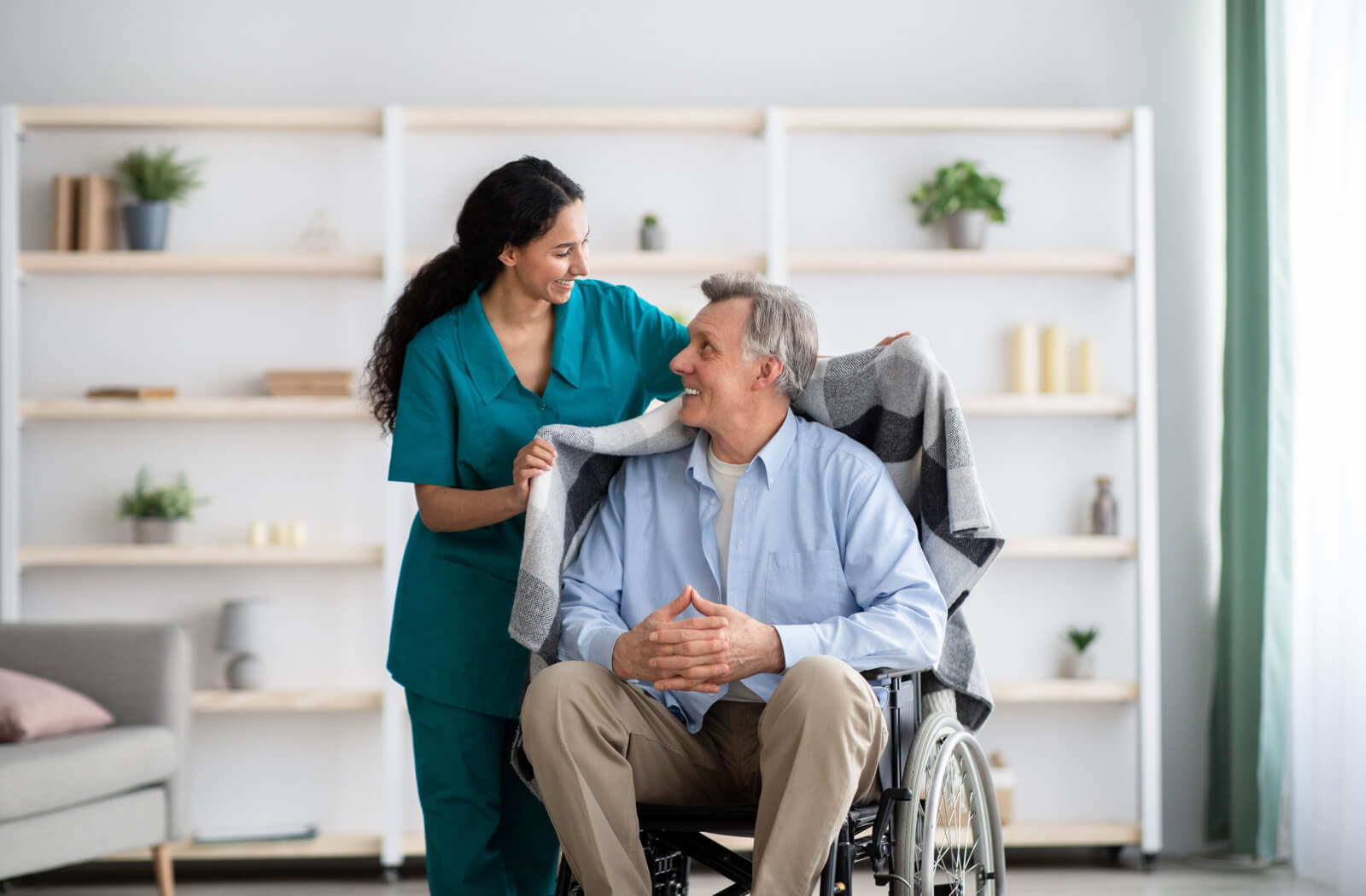 A female nurse helping a senior man in a wheelchair and covering him with a blanket.