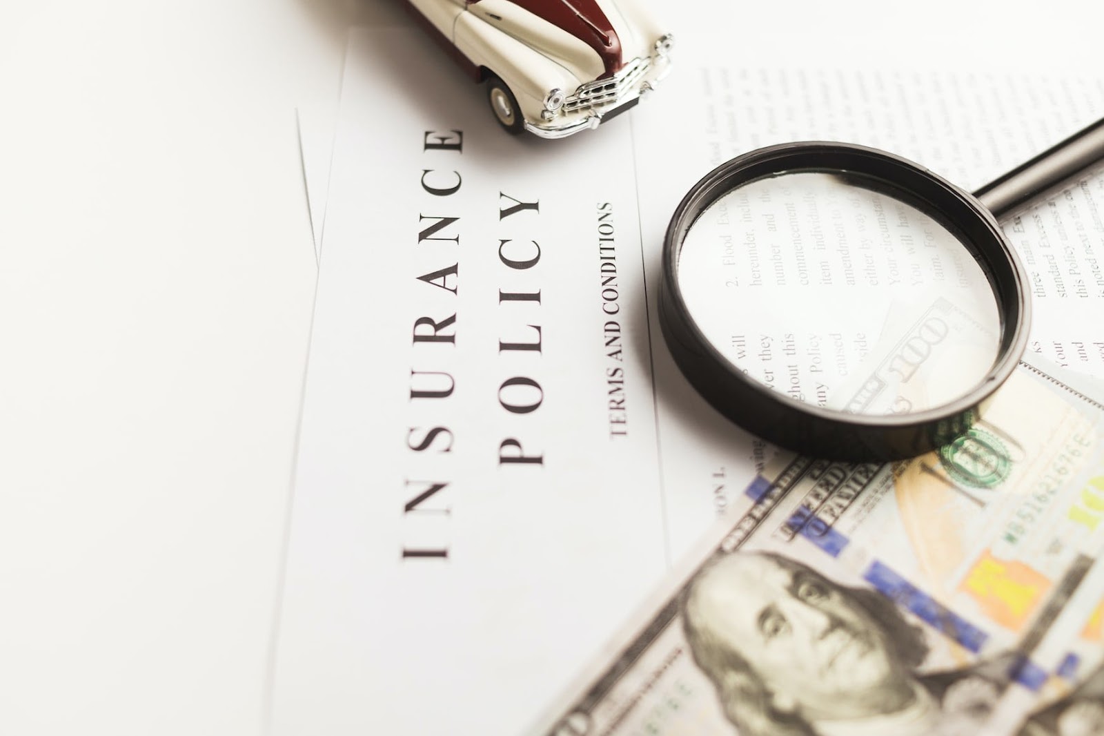 A printed copy of car insurance policy with a miniature car, magnifying glass, and a 100-dollar bill 