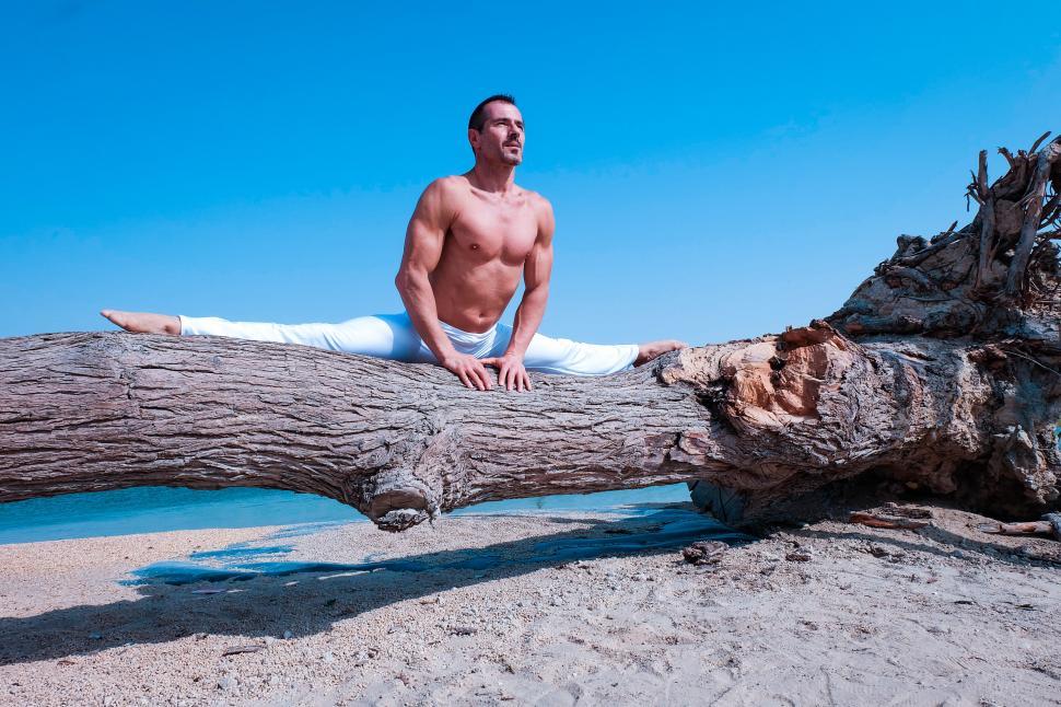 Man doing the splits on top of a fallen tree on the beach