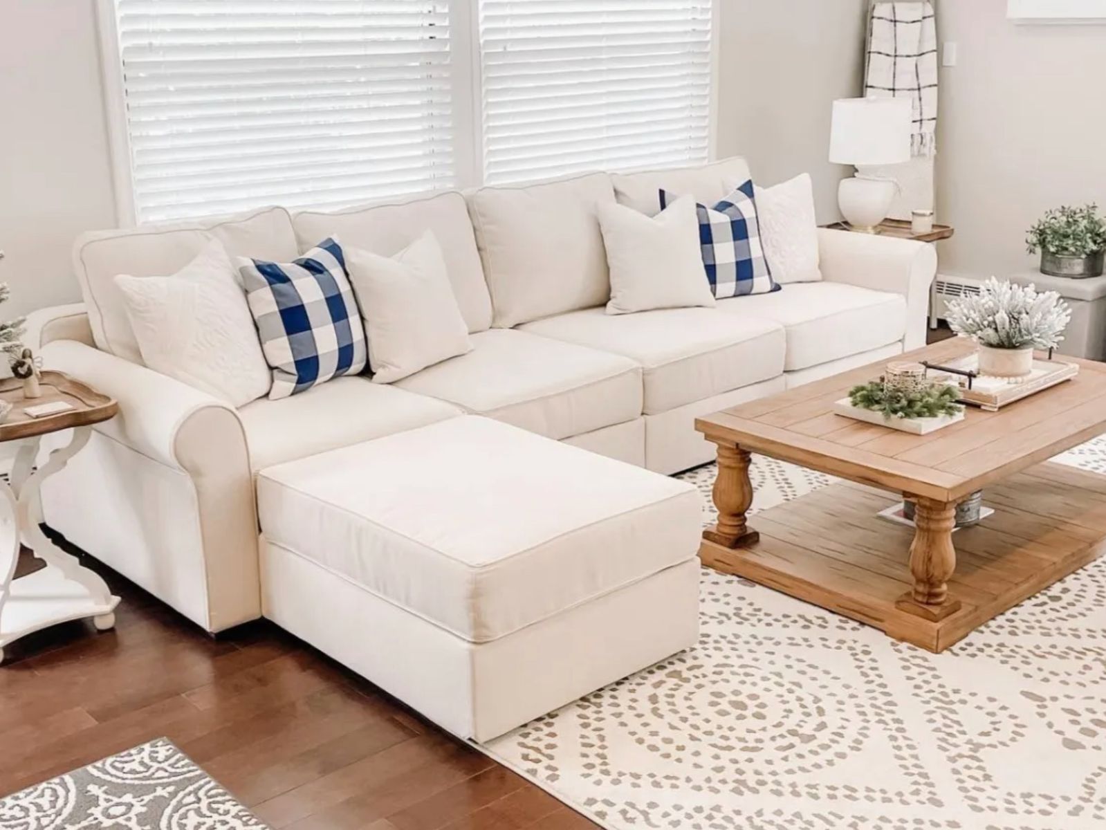 A cream chaise sectional from Lovesac is surrounded by traditional home furnishings including a farmhouse style coffee table, blue and white gingham throw pillows, and an abstract area rug.    